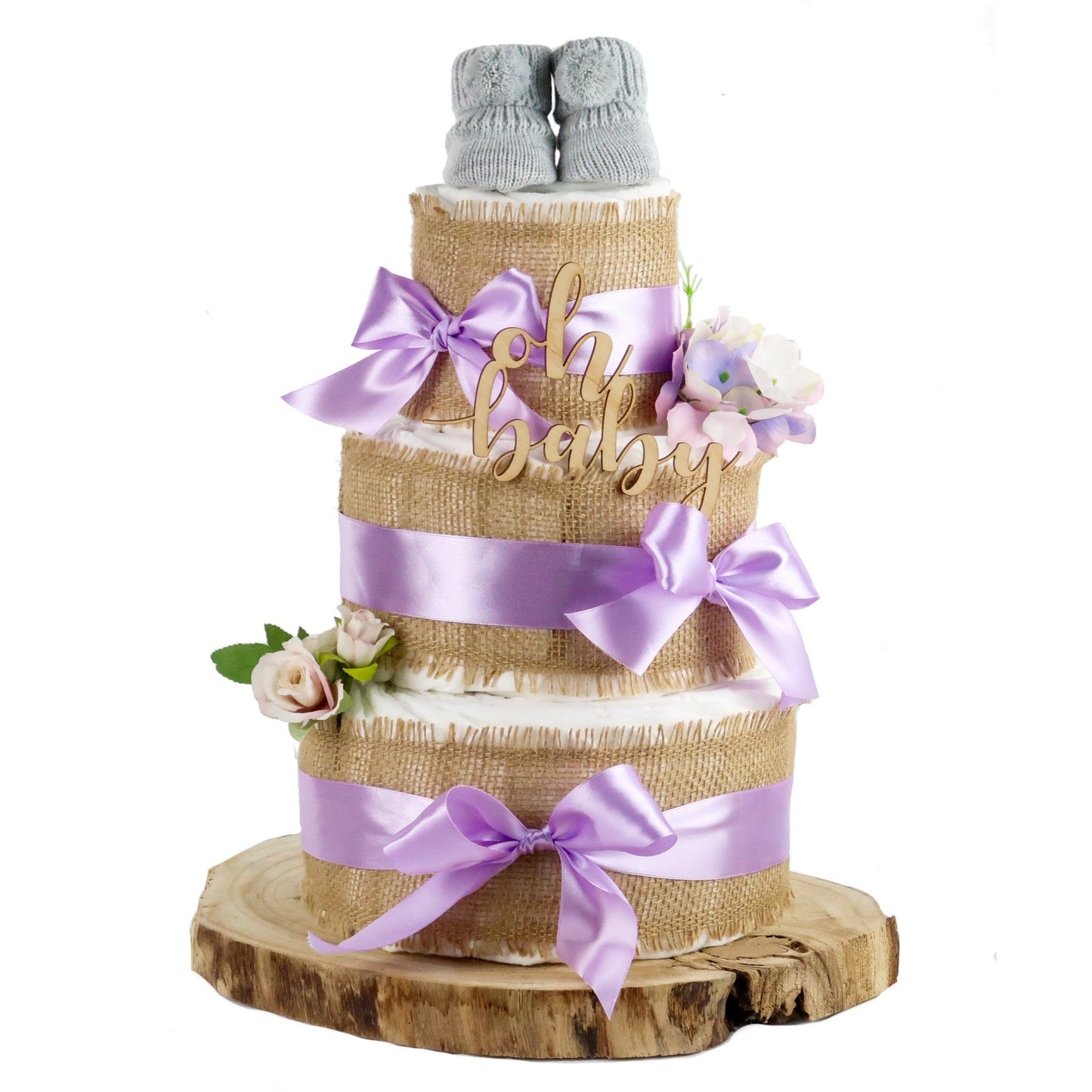 Three-tier diaper cake with burlap, lilac bows, flowers and grey baby booties displayed on a wooden slice