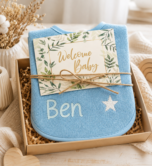 Blaues Lätzchen mit Namen Ben in einer Geschenkbox, verziert mit „Welcome Baby“-Karte und Juteband